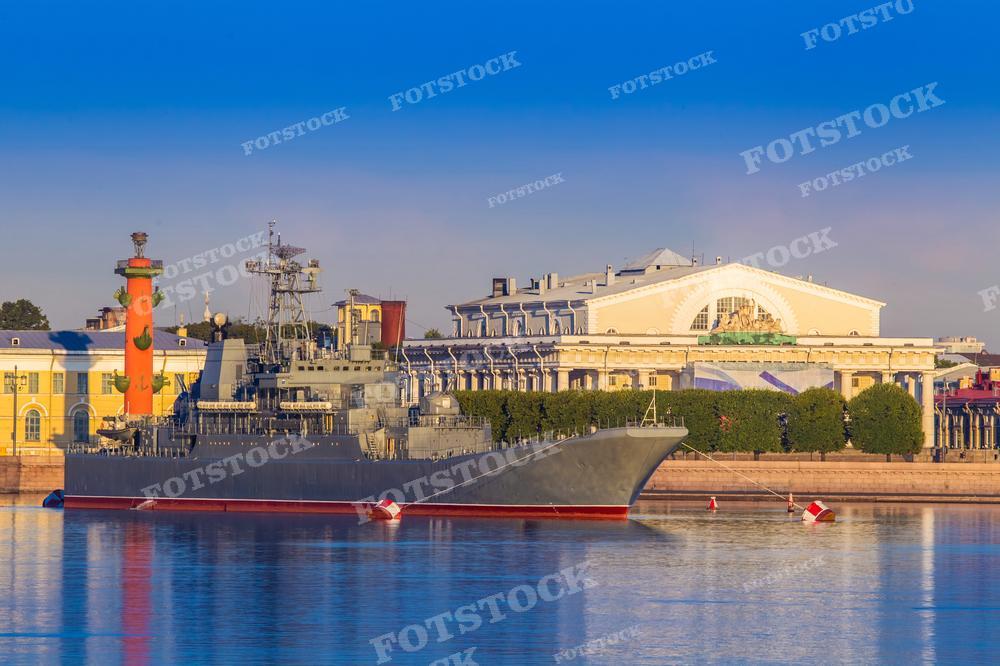 На фотографии запечатлен военный корабль на реке Нева в Санкт-Петербурге. На заднем плане расположены известные городские достопримечательности, в том числе Ростральная колонна и часть архитектуры Васильевского острова. Небо ясное, что указывает на раннее утро или поздний вечер с мягким светом. Эта фотография может быть использована для статей о военно-морском флоте, истории России, туристических публикациях о Санкт-Петербурге или как элемент образовательных и патриотических материалов.